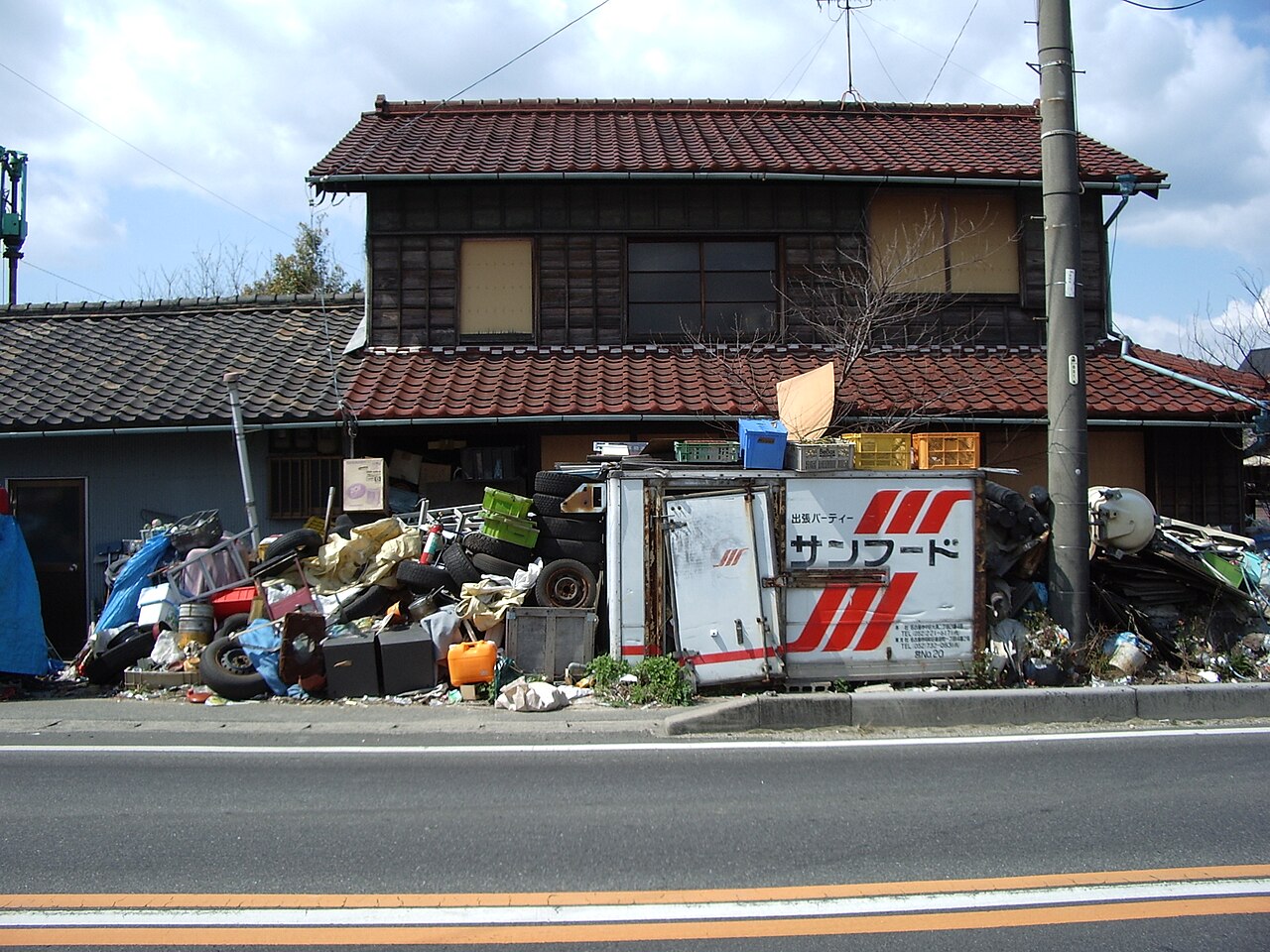 家財やタイヤ、荷台などが敷地の前面に積み上がった日本の戸建て住宅の外観